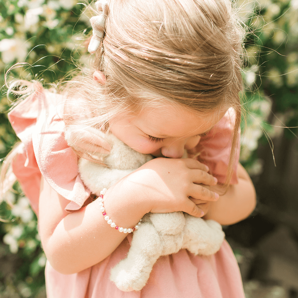 Little girl snuggling a fluffy bunny. She's wearing a pink ruffled dress with an adorable pearl and birthstone bracelet.